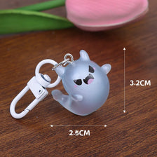 A small, light gray ghost charm with black eyes is attached to a white keyring, shown on a wooden surface.