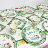 A green and white dinosaur party disposable tableware set with plates, cups, and a banner on a white tablecloth.