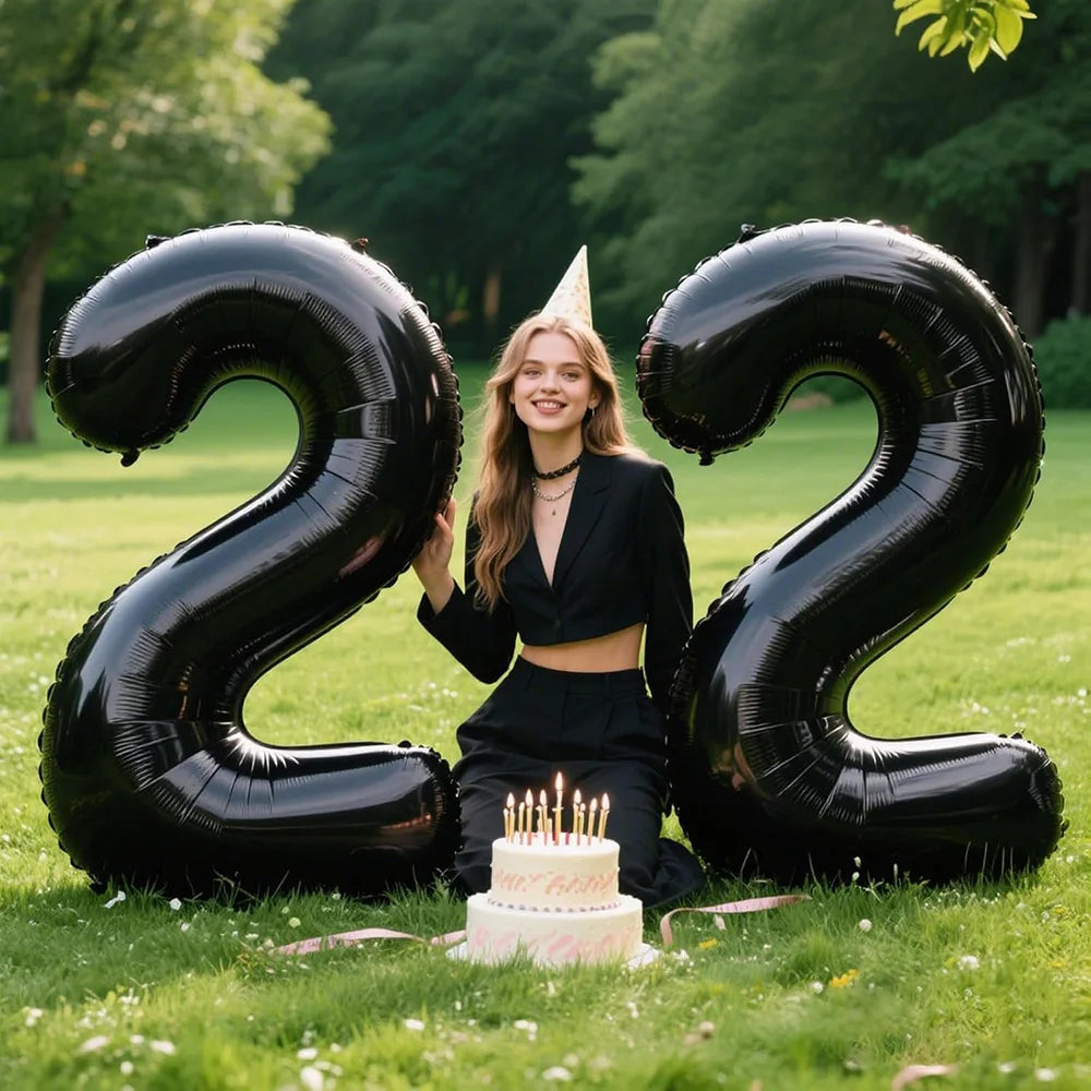 A woman is posing with large black foil number balloons spelling out 22 in a grassy outdoor setting.