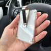 A clear acrylic visor clip decoration with a silver metal clip is held in a hand inside a car interior.