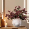 A faux branch of artificial nandina berry stem with dusty plum leaves displayed in a white vase next to candles.