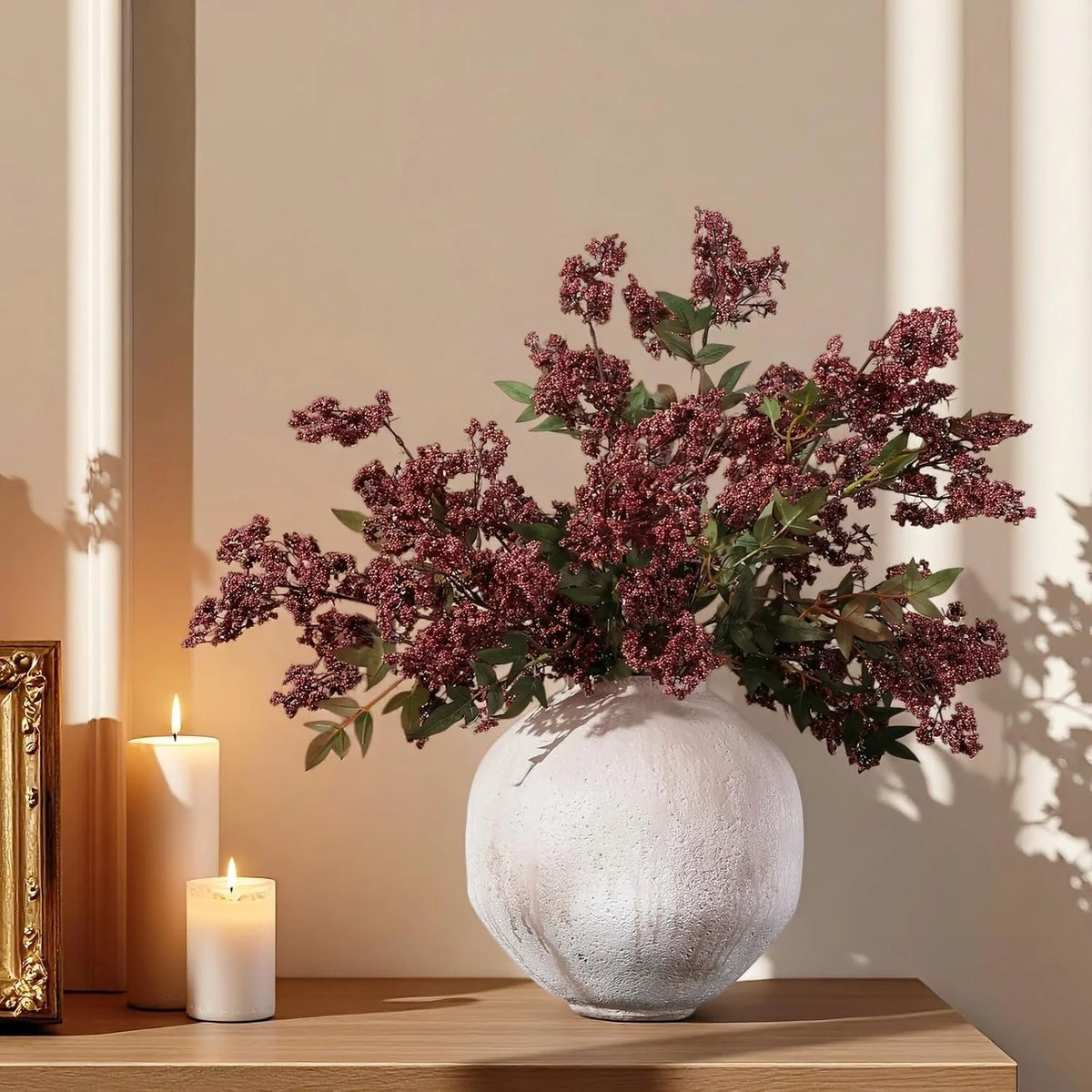 A faux branch of artificial nandina berry stem with dusty plum leaves displayed in a white vase next to candles.