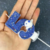 A hand holds a blue glitter number candle shaped like the number 2, featuring a white planet design.
