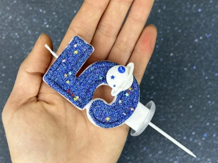 A hand holds a blue glitter number candle shaped like the number 2, featuring a white planet design.