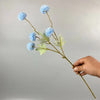 Una mano sostiene un tallo de flores artificiales de diente de león azules con hojas verdes contra un fondo gris claro.