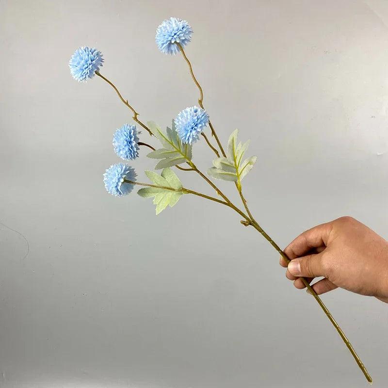 Una mano sostiene un tallo de flores artificiales de diente de león azules con hojas verdes contra un fondo gris claro.