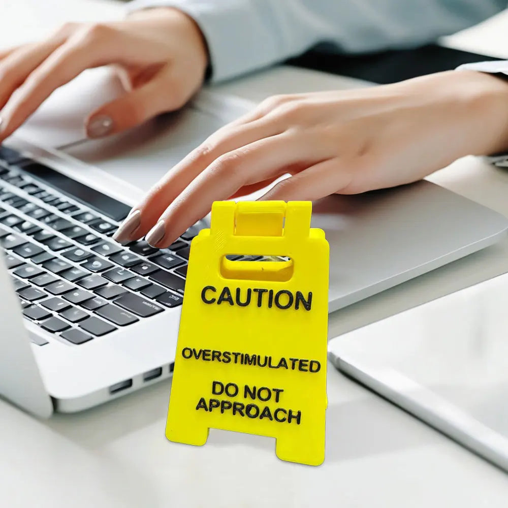 A yellow desk sign with black text warning caution and not to approach for overstimulated days placed on a laptop.
