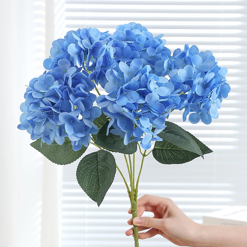 A cluster of artificial blue hydrangea silk flowers with green leaves being held by a hand.