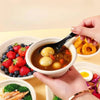 A hand is holding a bowl of soup with yellow ingredients and vegetables, surrounded by fresh berries and other food items.