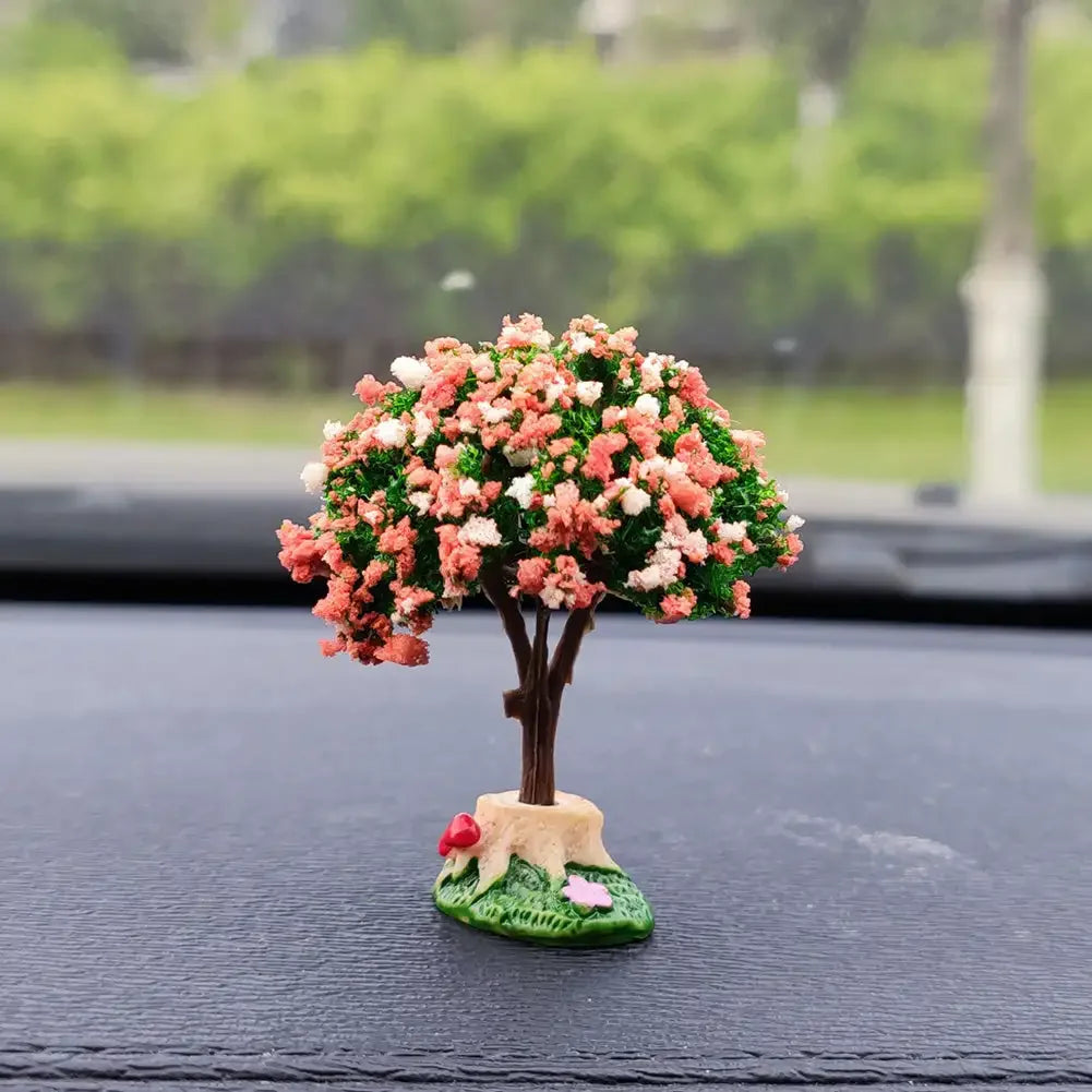 A miniature tree ornament featuring pink and white flowers displayed on a black car dashboard.