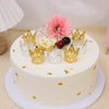 A white cake decorated with gold crown toppers, pink flowers, and dark berries for a celebration.