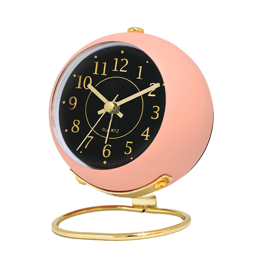 A minimalist gold-framed analog alarm clock with a pink face and black dial displaying the time.