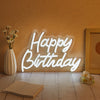 A glowing white neon sign spelling out Happy Birthday on a textured orange wall with dried flowers and wooden elements.