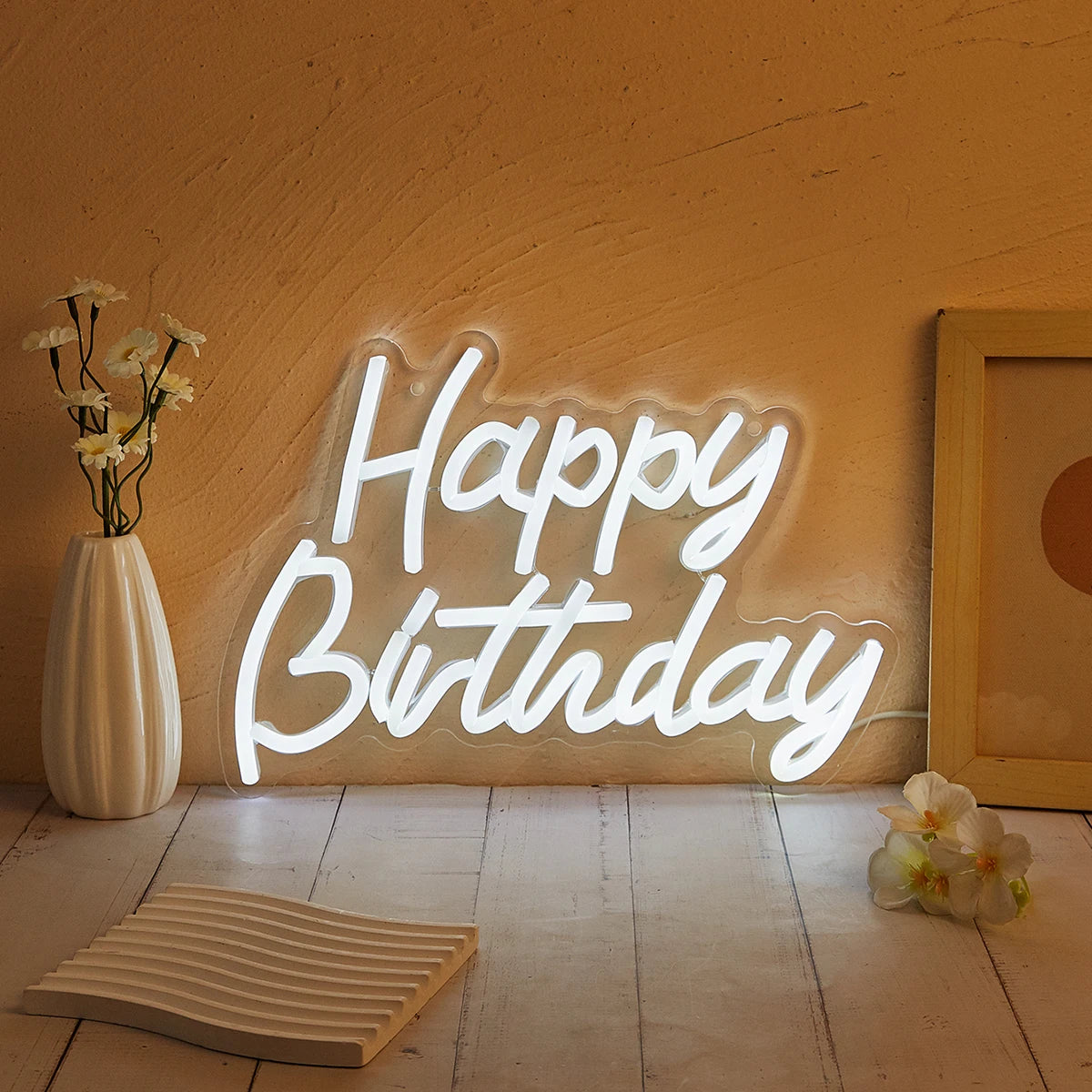 A glowing white neon sign spelling out Happy Birthday on a textured orange wall with dried flowers and wooden elements.