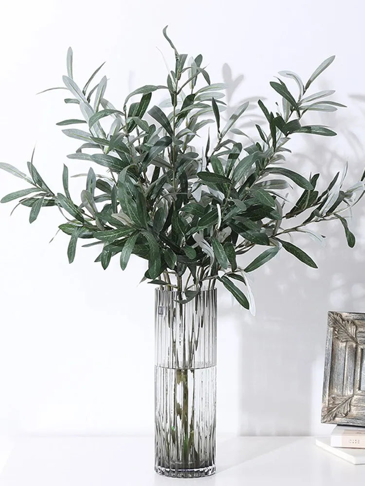 Artificial olive branch with green leaves and small fruits displayed in a clear glass vase.