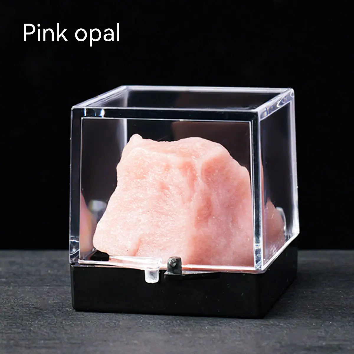 Pink opal rough stone specimen displayed in a clear box on a dark surface.