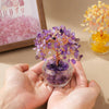 A hand holds a purple crystal money tree ornament with orange roots and a glass bowl, next to a yellow crystal arrangement.