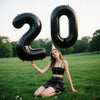A young woman sitting in a grassy field holding a large black foil number balloon spelling '20'