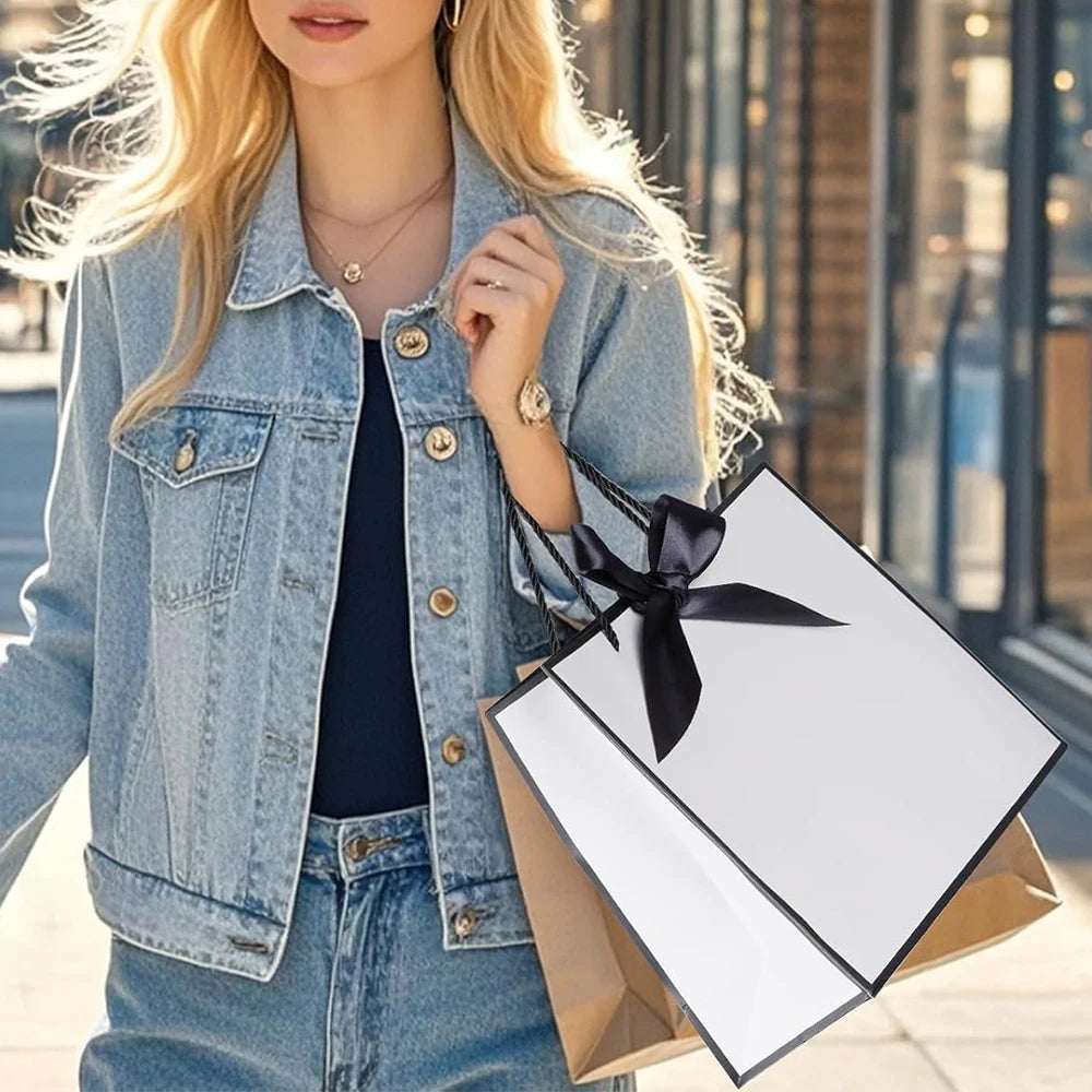A blonde woman wearing a denim jacket holds an elegant black and white gift bag with a black bow ribbon.