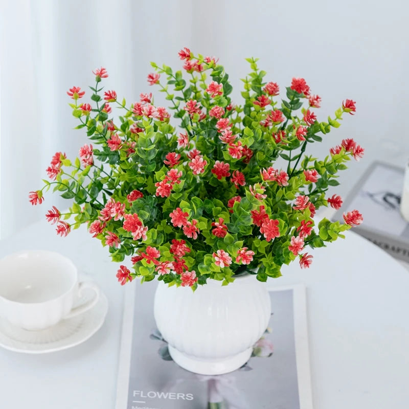 A bouquet of artificial eucalyptus branches with red flowers in a white vase on a white table.