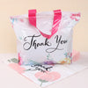 A white floral thank you gift bag with pink handles and black script writing on the front