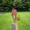 A rustic wrought iron cat ornament with a curved tail and pointed ears stands on a wooden post in a grassy yard.
