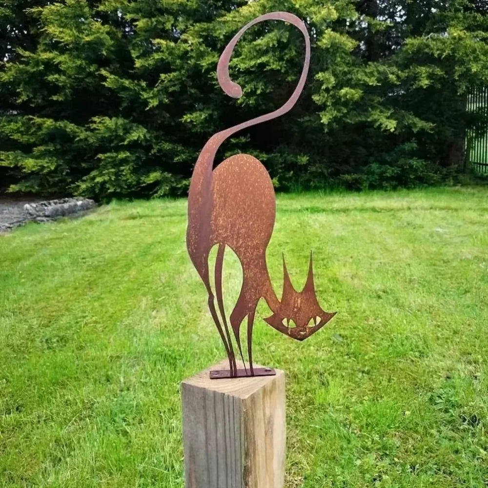 A rustic wrought iron cat ornament with a curved tail and pointed ears stands on a wooden post in a grassy yard.
