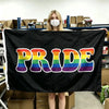 A person holds up a black banner displaying the word PRIDE in a rainbow flag design.
