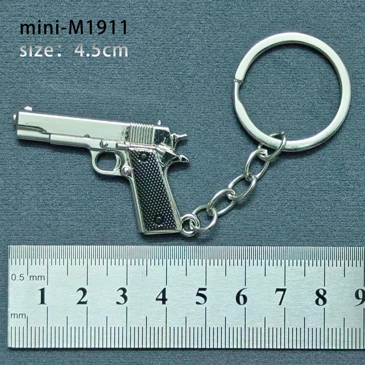 A silver miniature replica of a pistol is attached to a keyring, measured against a millimeter ruler.