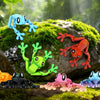 A collection of colorful, articulated 3D printed frog magnets in various shades of blue, green, and orange resting on mossy r