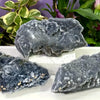 A cluster of dark gray natural zinc crystals displayed on a white surface with green foliage in the background.