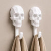 A pair of white 3D skull wall hooks displaying a textured, modern design, mounted on a beige wall above towels.