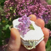 A hand holds a mini metal flower press kit with dried purple lilac flowers against a blurred background of pink blossoms.