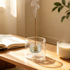 A clear glass incense burner with a dark stick emitting smoke on a wooden table.