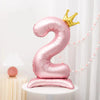 A pink foil number balloon shaped like the number 2 with a gold crown topper displayed on a white stand.