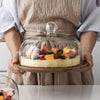 A person holds a light yellow cake on a wooden tray covered by a clear glass dome.