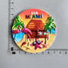 A circular magnet featuring a tropical scene with flamingos, palm trees, and a beach under the USA Miami title.