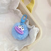 A fluffy blue plush sausage-mouth keychain with purple lips and a blue clasp is shown inside white tissue paper.