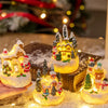 A hand holds a set of glowing Christmas gingerbread house resin ornaments resting on a snowy surface.