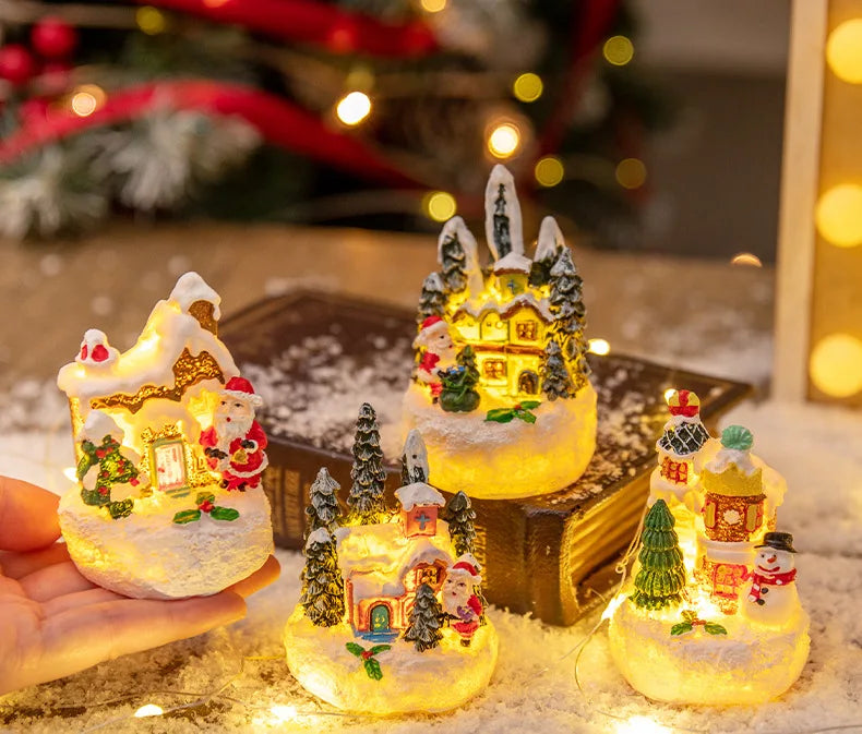 A hand holds a set of glowing Christmas gingerbread house resin ornaments resting on a snowy surface.