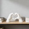 A white heart shape sculpture formed by two hands resting on a wooden shelf.