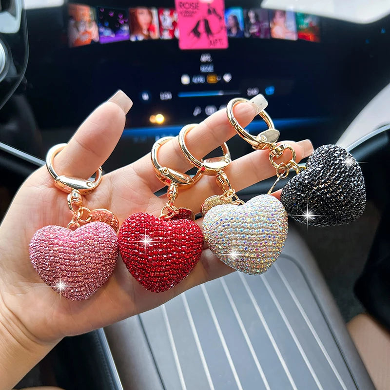 A hand displays five heart-shaped crystal keychains in shades of pink, red, gold, and black.