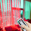 A hand holds a remote control for USB-powered LED curtain lights with pink and red lights illuminating a window.