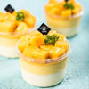 A serving of yellow custard or mousse in a clear plastic bowl with a lid, set against a light blue background.