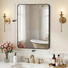 A modern wall mirror with a black metal frame reflects a bathroom interior and is flanked by brass sconces.