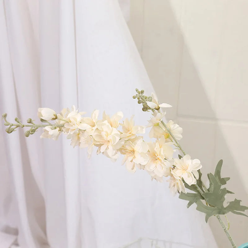 Un délicat jet de fleurs de delphinium blanches artificielles avec du feuillage vert sur fond blanc.
