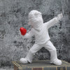 A white resin sculpture of a figure throwing a red heart against a textured gray background.