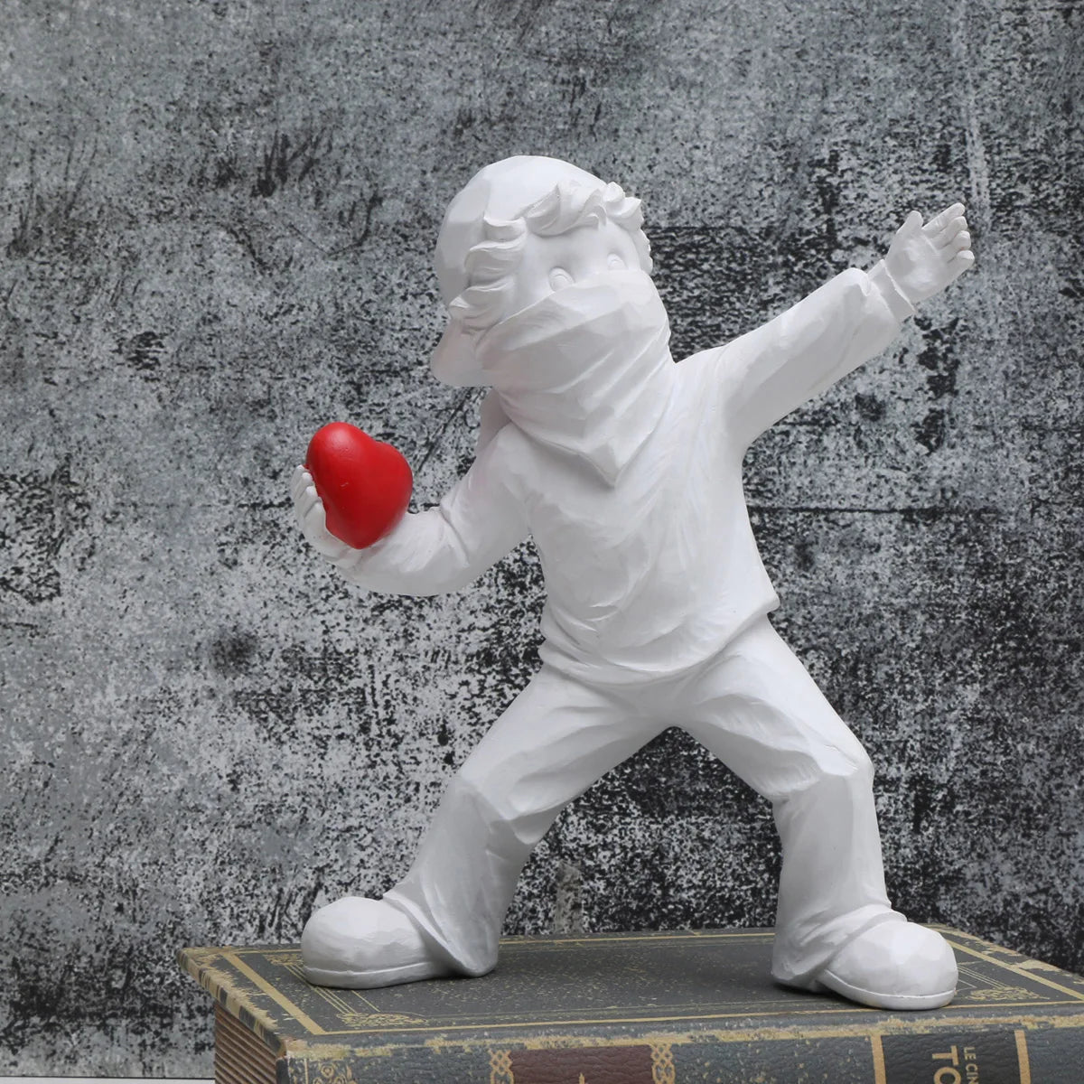 A white resin sculpture of a figure throwing a red heart against a textured gray background.