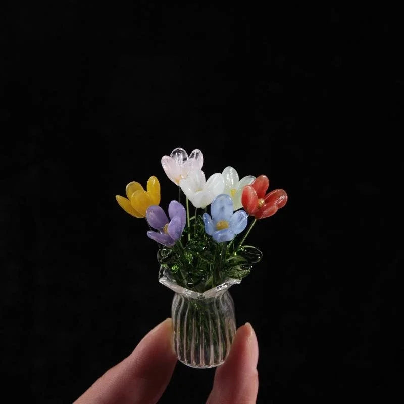 A hand holds a small glass vase filled with colorful glass flower sculptures.