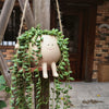 A light beige resin succulent planter with a smiling face is hanging from a wooden structure, surrounded by lush green vines.